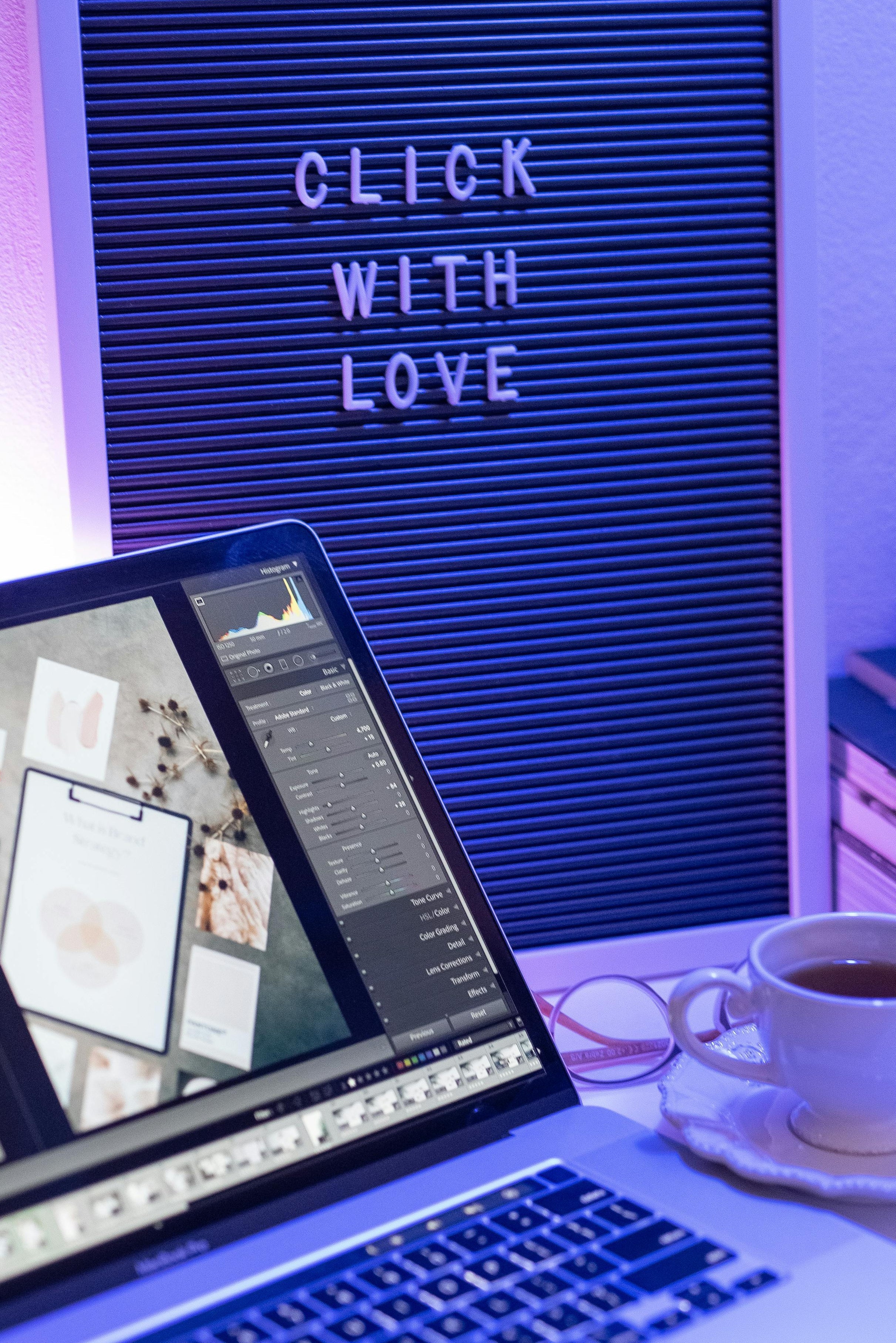 Laptop on a desk with a coffee cup and letter board displaying 'Click with Love' in a room with purple lighting.